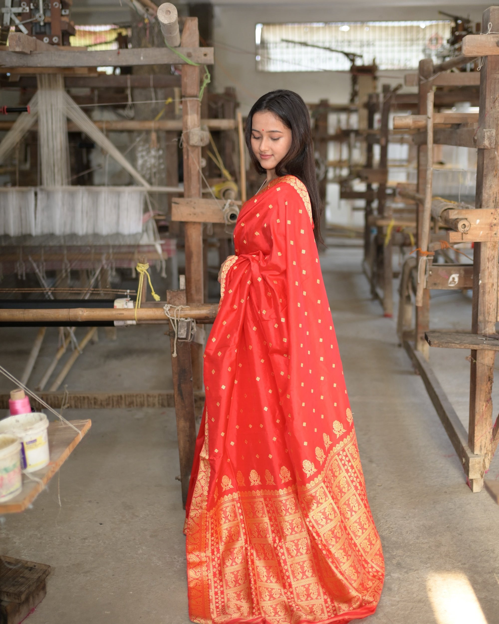 Turned three-quarters away between the handlooms, eyes lowered: the red drape full length and the golden panels of the pallu gathered at the hem.