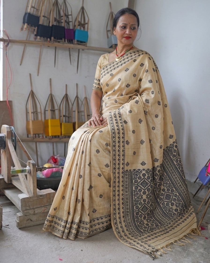 Seated beside a wall of bamboo reels wound with coloured yarn, the whole saree in view: small black motifs over a cream body, black border bands across the hem of the pleats, and the pallu falling from the shoulder to the floor in a dense black panel of diamond medallions, ending in a fringe.