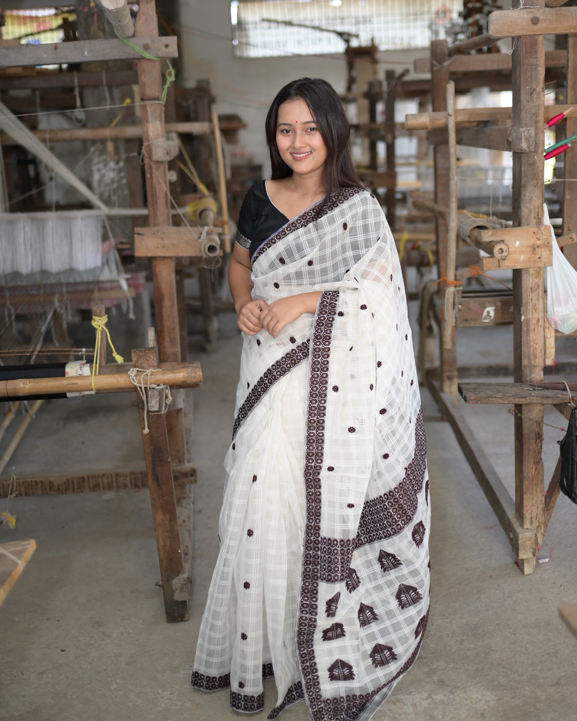 Standing among wooden handlooms in a weaving hall, smiling, hands together at the waist: the white check saree full length, small maroon flowers across the body, the maroon border down the pleats, and the pallu falling in front with rows of large peaked motifs.