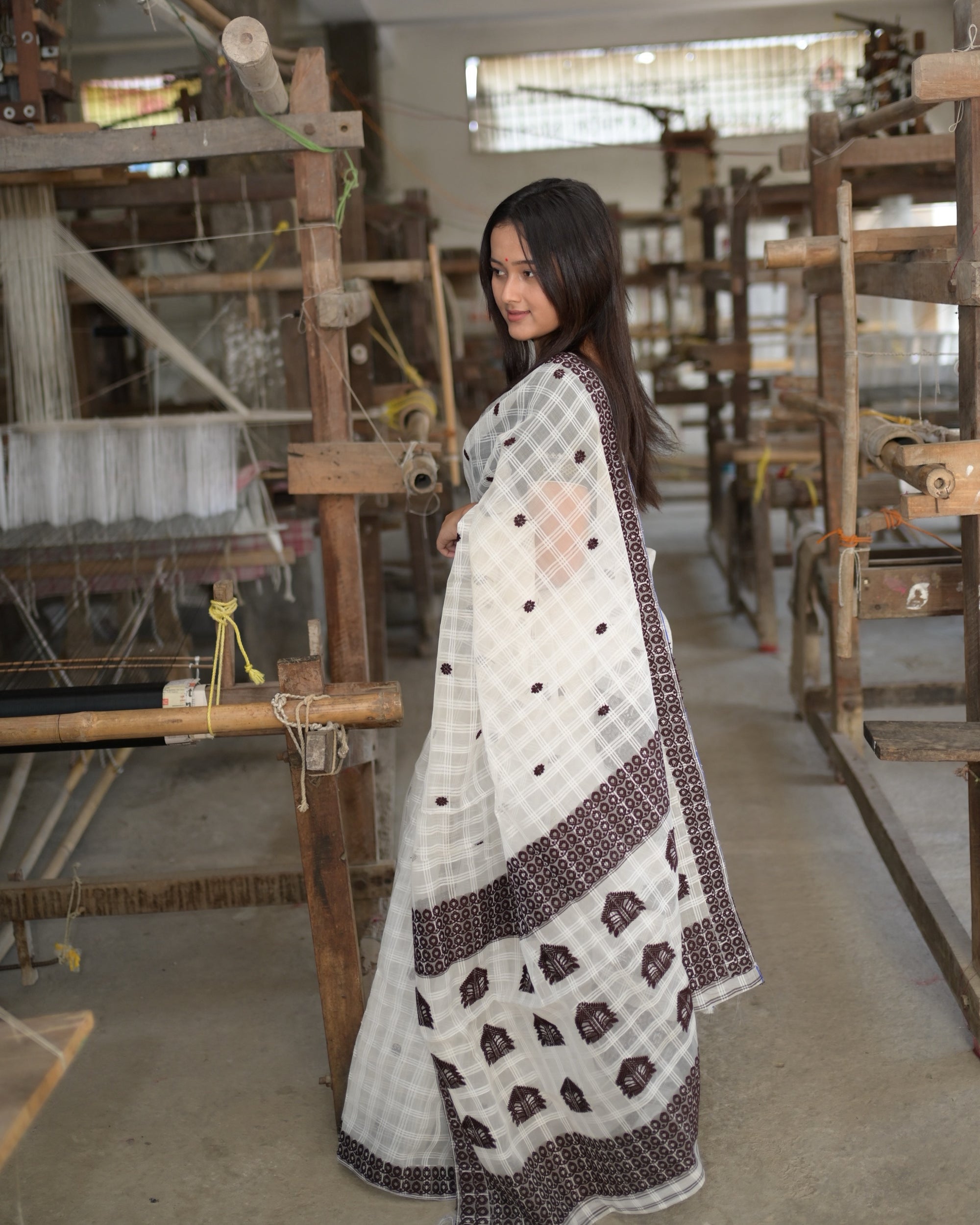 Turned to one side among the looms, looking back over her shoulder: the pallu falling to the hem with its maroon bands and large peaked motifs, small maroon flowers across the body.