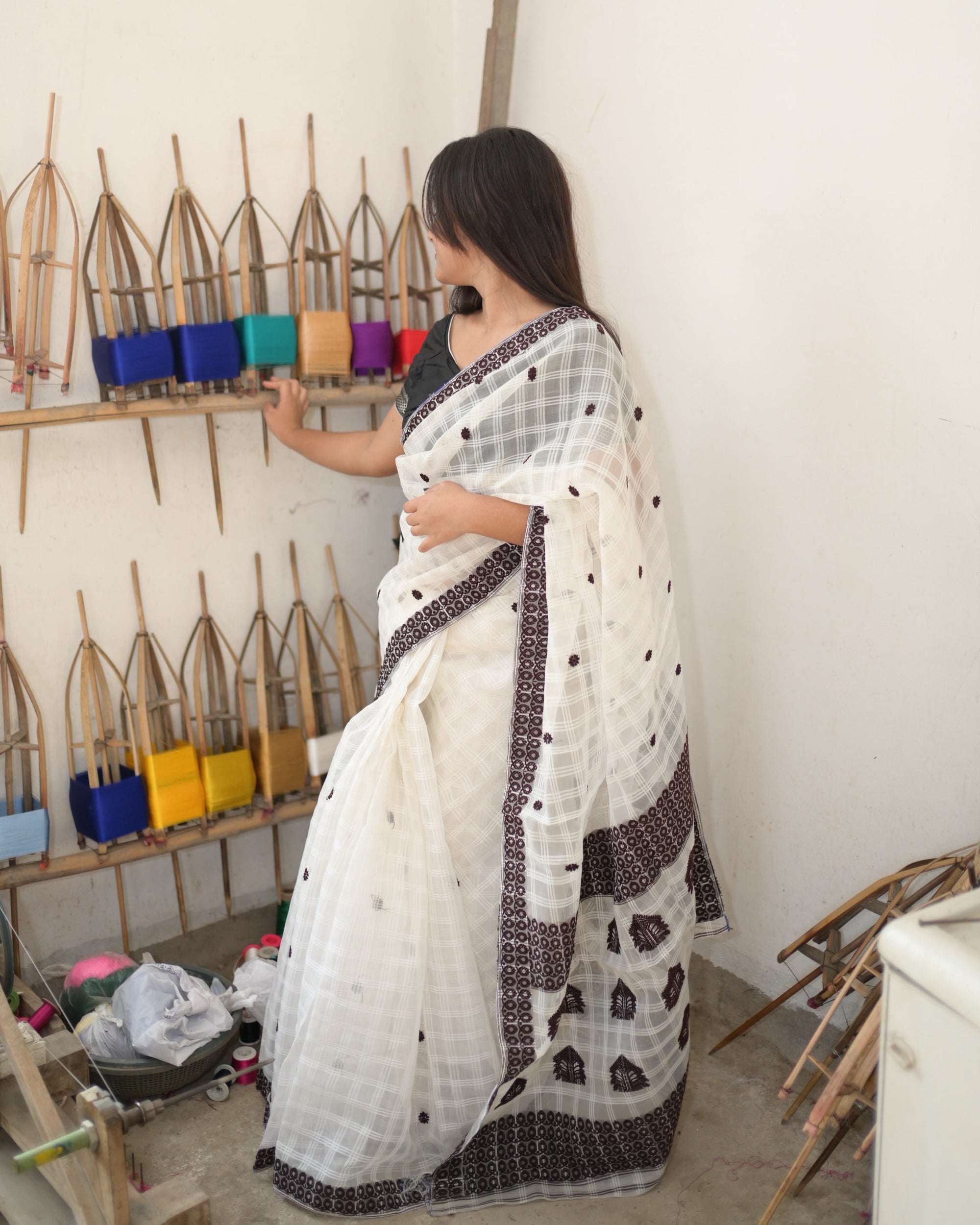 Beside a wall of bamboo reels wound with coloured yarn, one hand on the shelf: the white check saree from the side, the maroon border down the drape and the pallu’s large motifs near the hem.