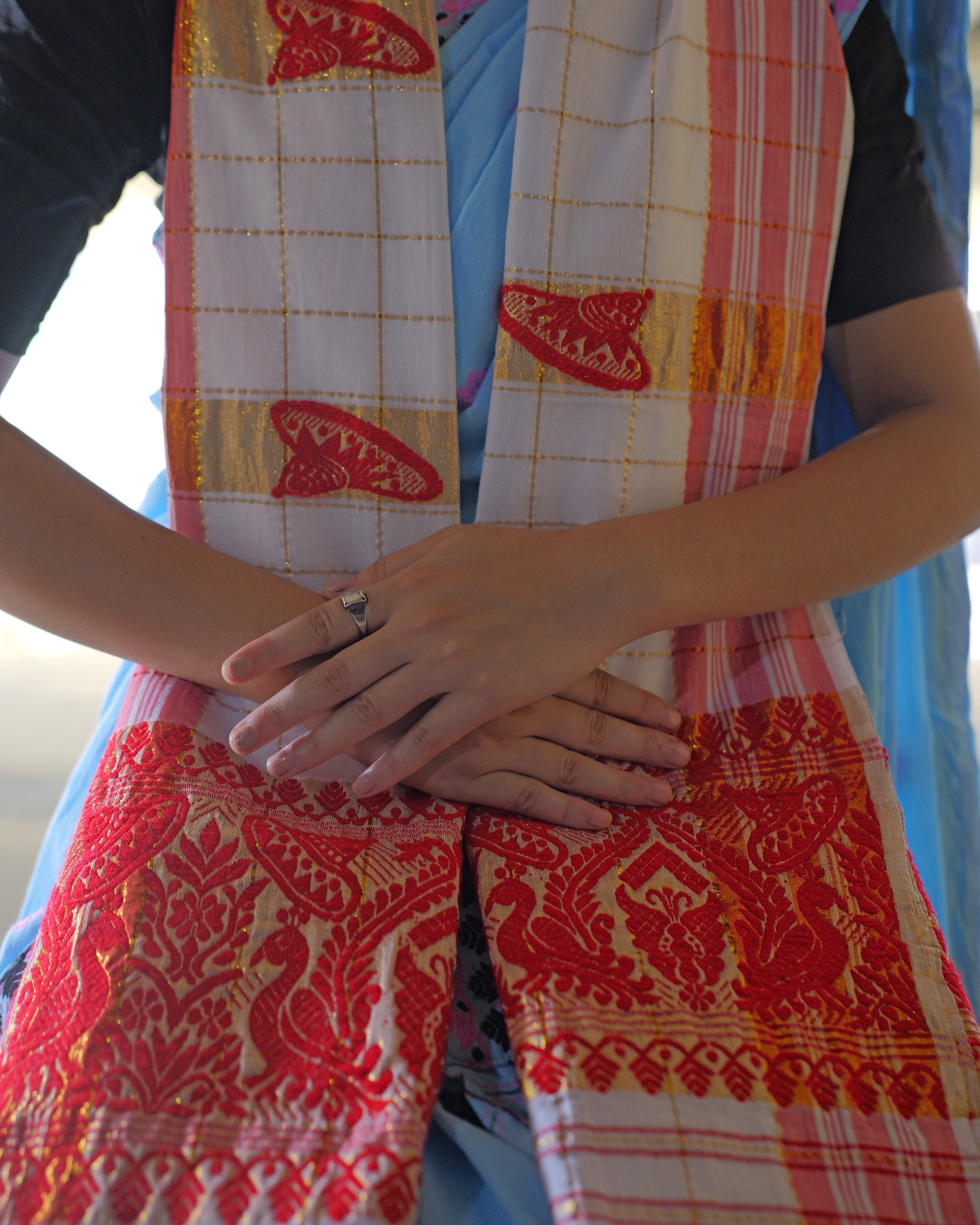 Hanging from the shoulders over a black blouse and a pale-blue sador, both ends falling down the front with the hands crossed over them at the waist, a silver ring on one finger. The white cloth is checked in fine gold lines, with red hat-shaped japi on broad gold bands, and each end is a dense red panel of birds and paisleys.