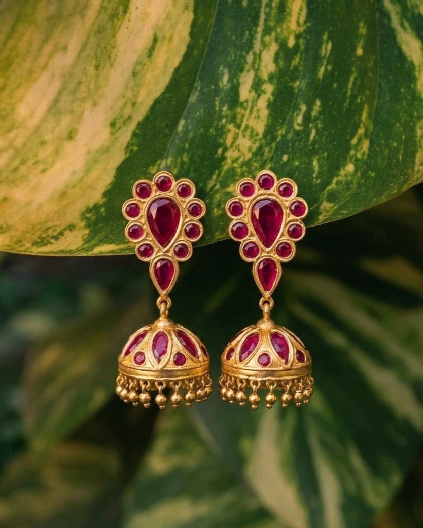 The pair hanging in front of a broad variegated leaf, green streaked with cream, more leaves blurred behind. Each earring is a rosette of red stones around a red teardrop, a second red teardrop below it, and a gold bell set with red stones and fringed with small gold beads.