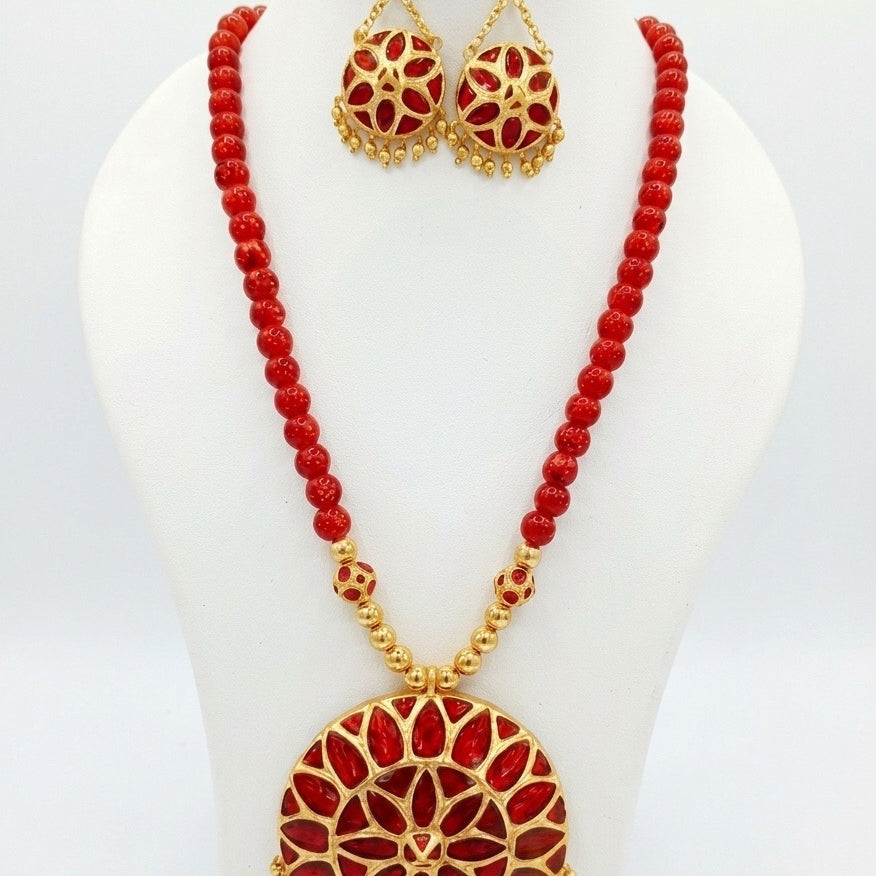 On a white display bust against a white ground: a large round gold medallion filled with red petal shapes in two rings, fringed with small gold beads along its lower edge, on a strand of round red beads finished with gold beads near the pendant. The earrings are smaller red discs hung from three-stone buds on fine chains.