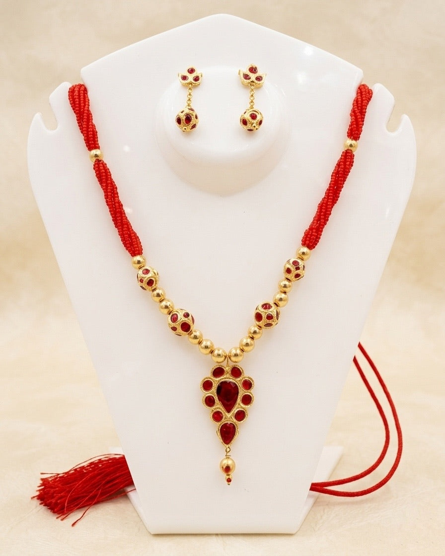 On a white acrylic necklace stand against a soft cream ground: a small gold teardrop pendant of red stones, a large red teardrop ringed by small ovals with a gold drop below, on plain gold beads and gold beads set with red stones that lead up to twisted strands of red seed beads. A red cord and tassel lie at the foot of the stand, and the studs, with stone-set gold ball drops, sit on the round platform at its top.