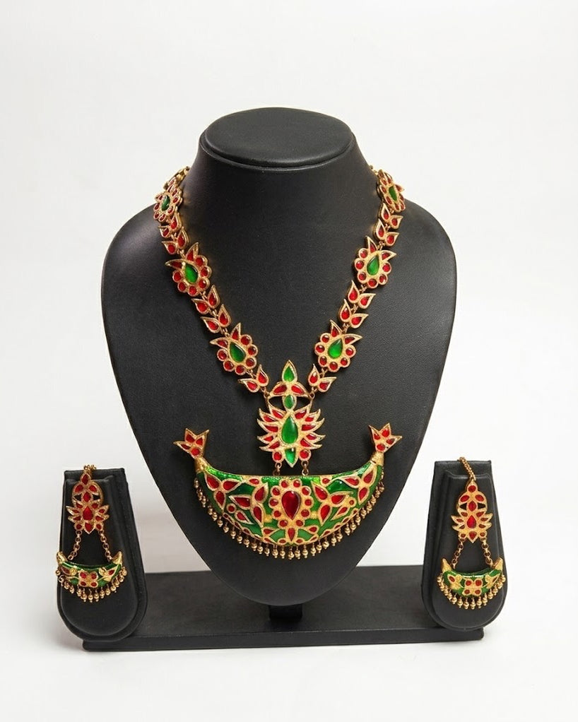 The necklace on a black display bust, its earrings on their own black stands either side, against a pale grey ground. A V of small medallions, each a green stone ringed with red, runs down to a red lotus and a crescent with a bright green face set with red stones, a bud at each tip and a fringe of gold drops. The earrings hang small green crescents from tops of red stones.