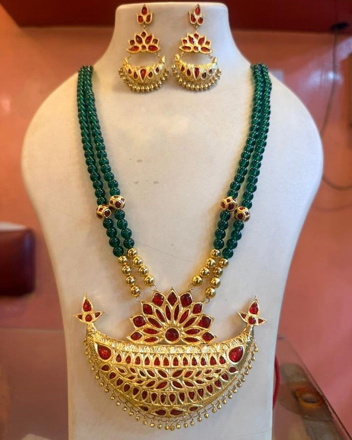 The necklace and earrings on an ivory display bust on a shop's glass counter, a pink wall behind. Two strands of dark green beads, ending in plain gold-toned beads, carry a broad gold-faced crescent set with rows of small red stones, a fan of red petals standing on its top edge and a fringe of gold drops. Three-tier earrings set with red hang at the neck.