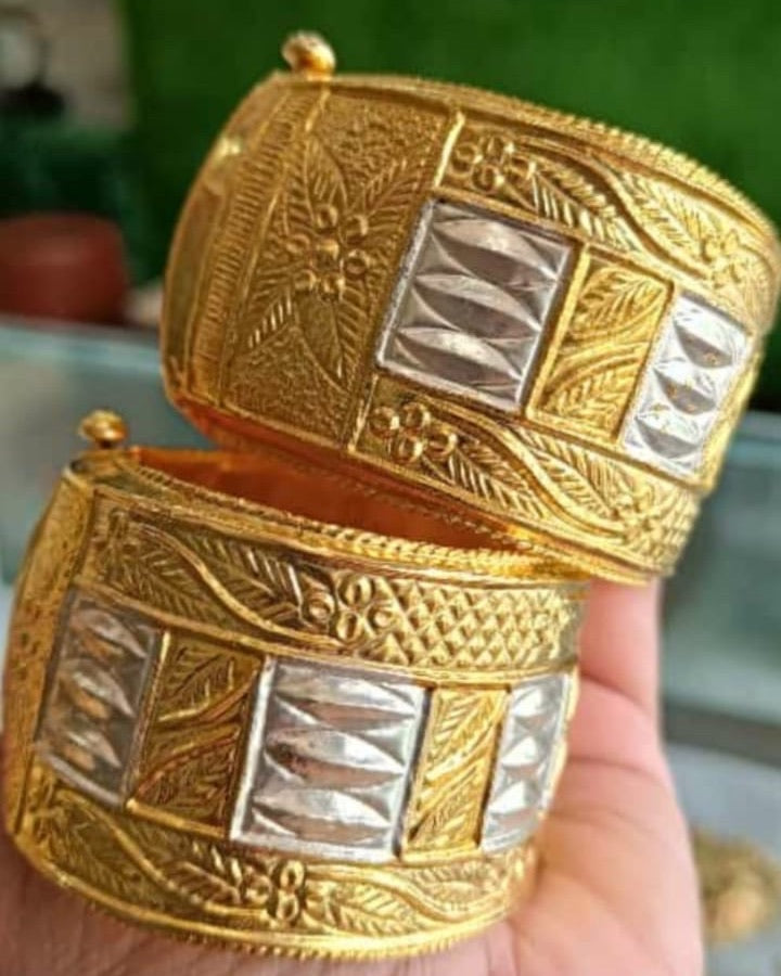 A hand holds the two bangles, one stacked on the other, against a blurred green wall and a glass counter. Each is a broad gold-toned cuff with a band of square silver-white panels, each raised in four stacked almond shapes, between gold panels engraved with leaves; vines and small dot-flowers run along the borders, and a ball-headed pin shows at the top edge.
