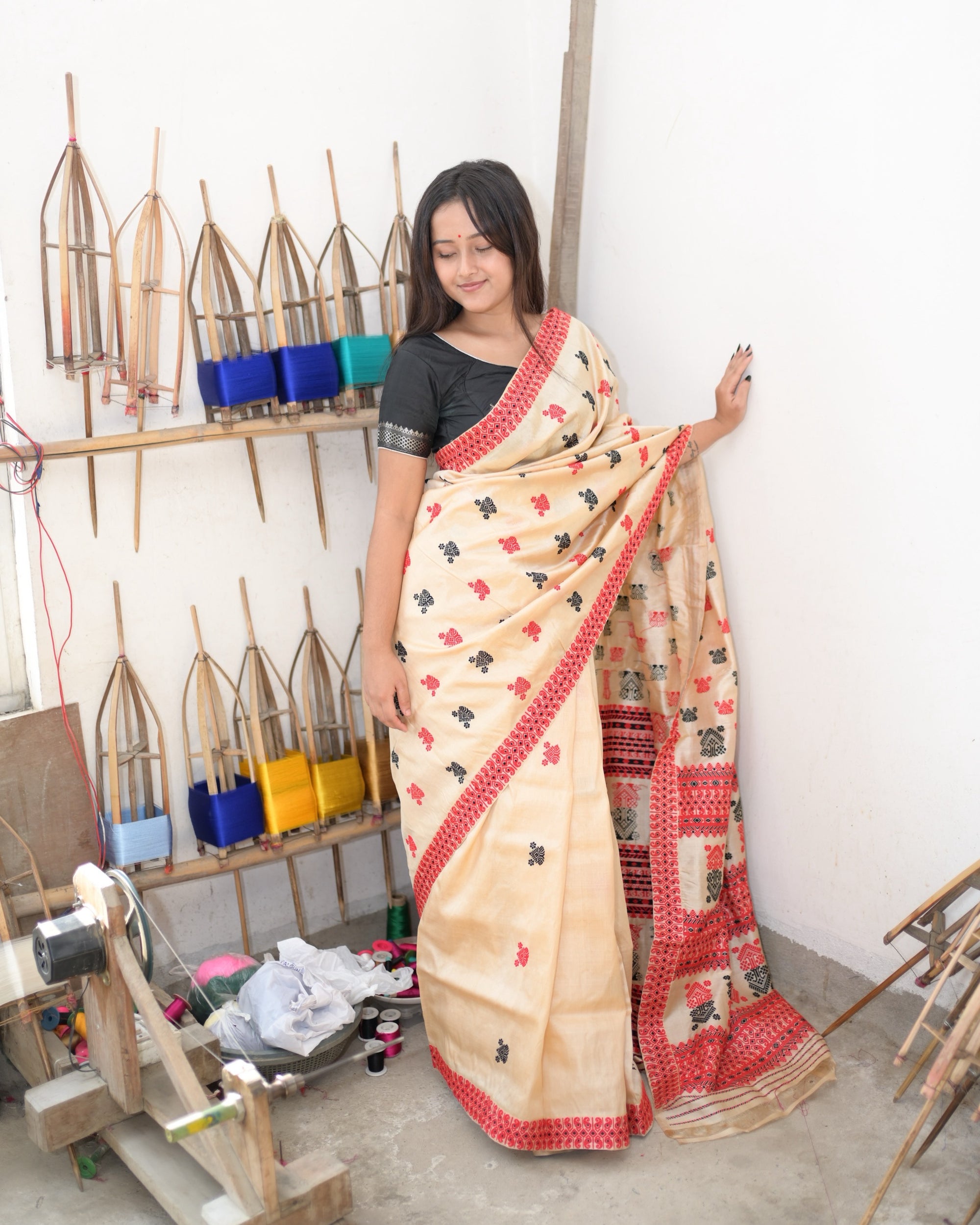 Standing beside a wall of bamboo reels wound with coloured yarn, one hand raised to the wall: the cream saree full length, small red and black buti across the body, the red scroll border down the pleats, and the pallu falling from the shoulder in red lattice bands and paired medallions.