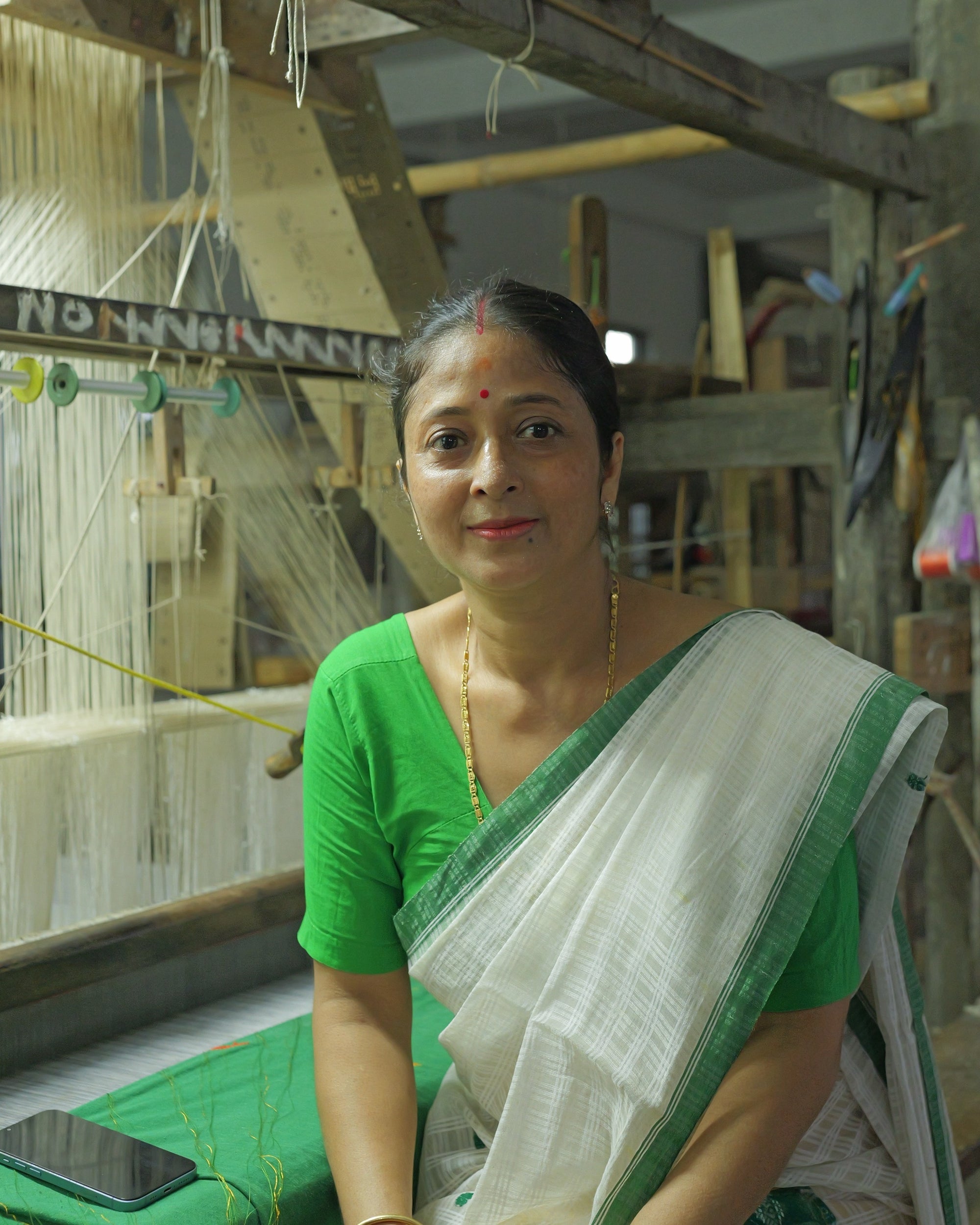 Piju Moni Das seated at a loom in a green blouse and a white drape with a green border, smiling at the camera, white warp threads strung on the loom beside her.