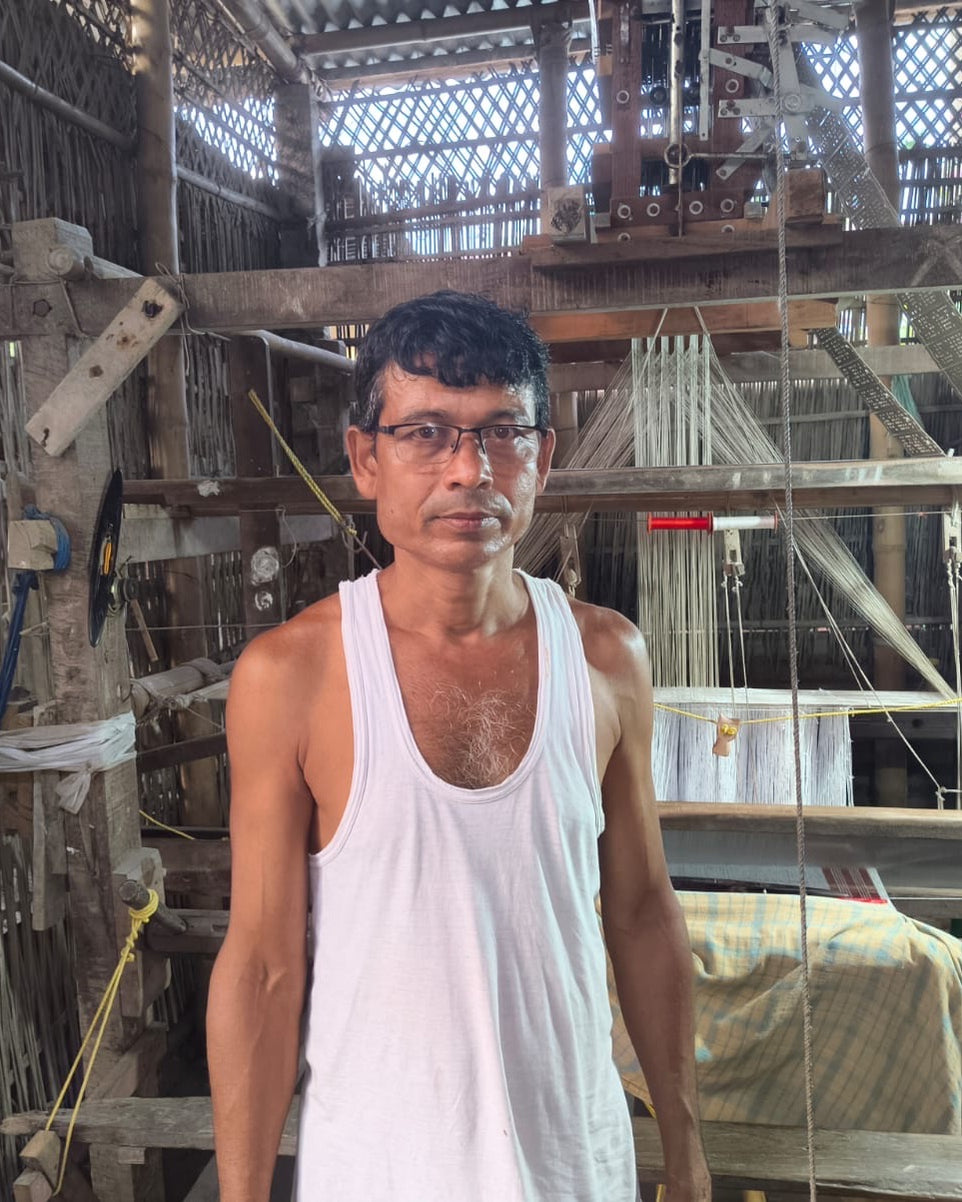 Ratan Baishya standing in a bamboo-walled loom shed in a white vest and glasses, facing the camera, a loom with a punched-card jacquard head and a hanging chain of cards behind him.