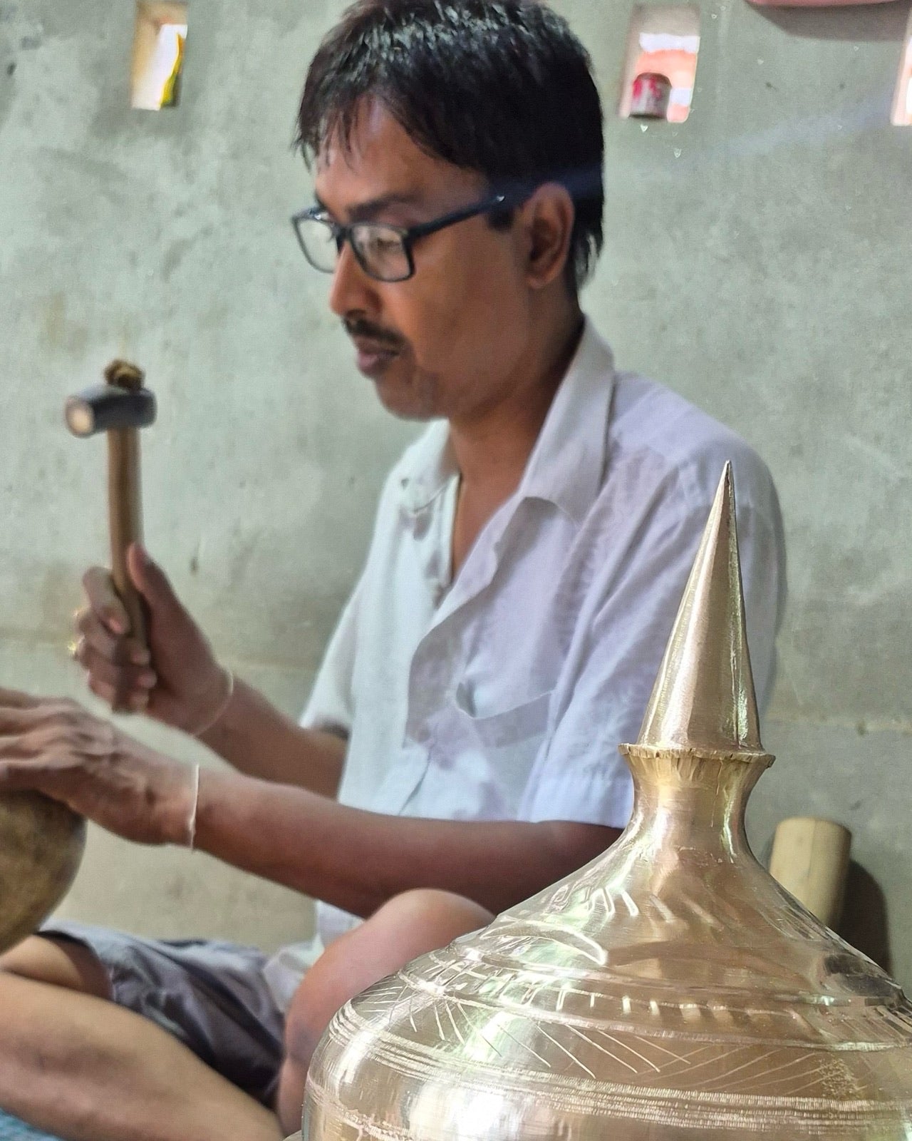 Haridas Das seated cross-legged on the workshop floor in glasses and a white shirt, hammering a rounded metal dome held over an iron stake, the polished lid and spire of a finished xorai in sharp focus in front of him.