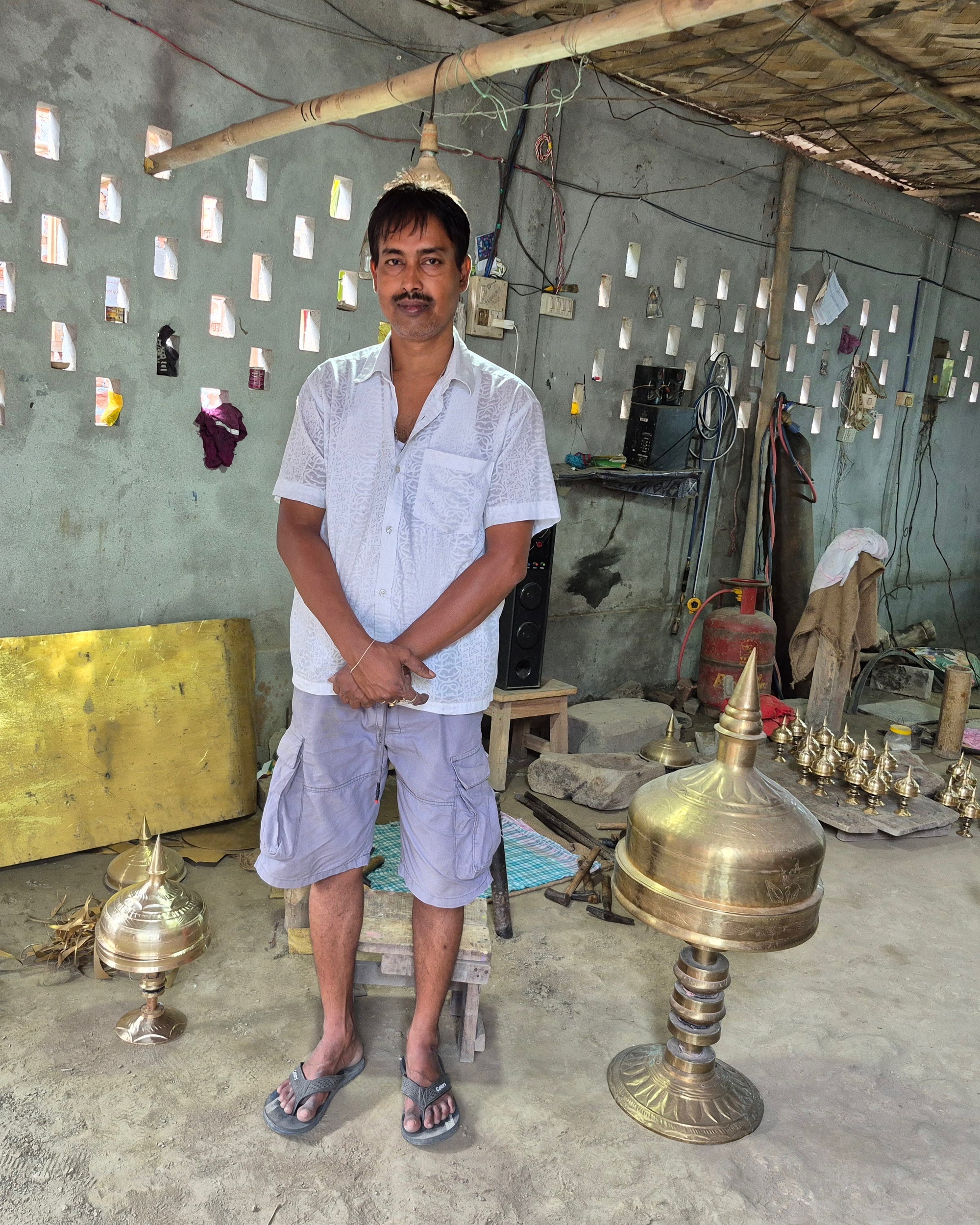 Haridas Das standing in a metal workshop in a white shirt and grey shorts, hands clasped, facing the camera, finished xorai on the earth floor beside him and a sheet of yellow metal leaning on the wall behind.