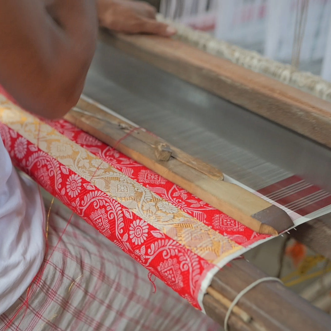 Over a weaver's shoulder at a loom: below the arm, cloth woven in red and gold-coloured bands of flowers, a wooden temple lying across it.