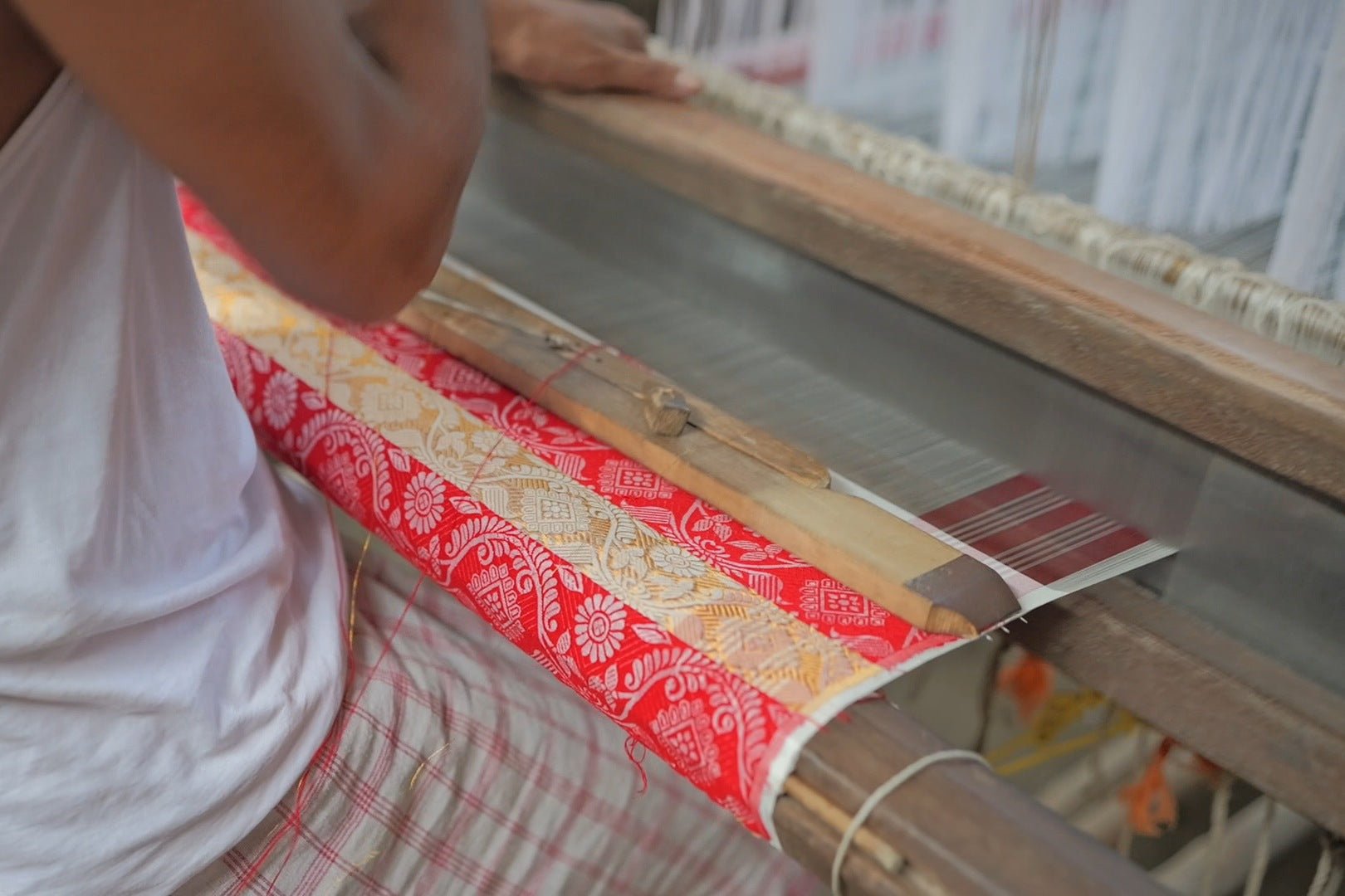 Over a weaver’s shoulder at a loom: below the arm, cloth woven in red and gold-coloured bands of flowers, a wooden temple lying across it.