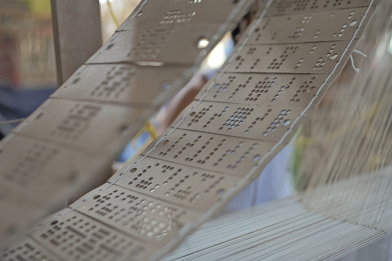 A long chain of laced punched cards, grey-brown and pierced with groups of round holes, curving down beside a loom, white cords and warp behind.