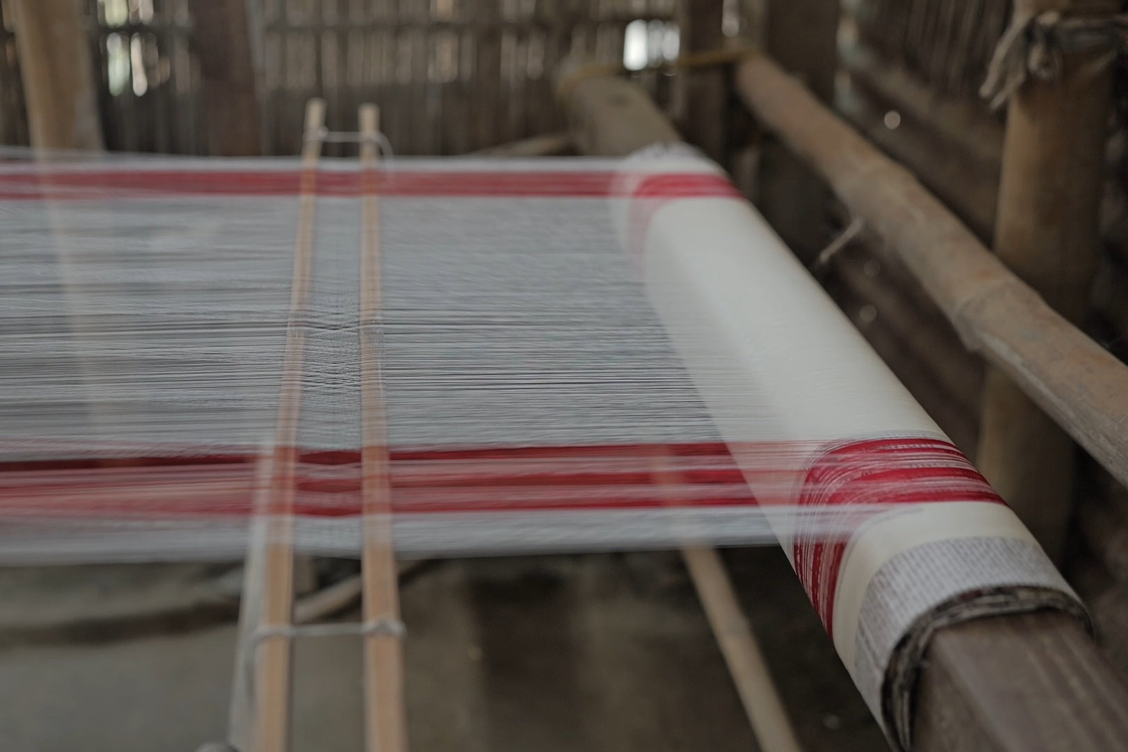A white warp with groups of red threads at both edges, wound thick on a loom’s back beam, two tied lease rods crossing it, split-bamboo walls behind.