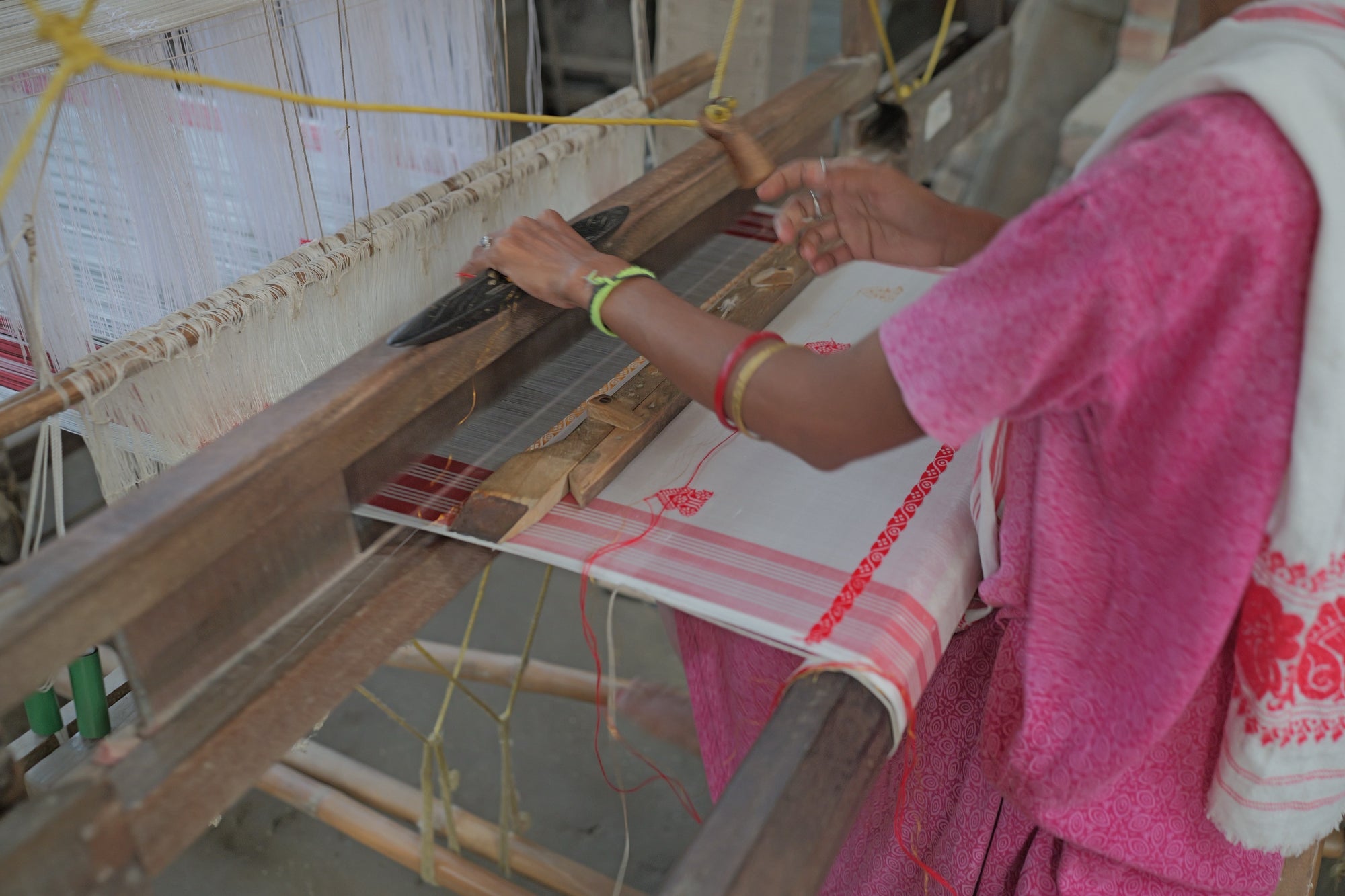 Hands at a wooden loom: the left grips a dark boat shuttle on the batten, the right is raised over white cloth with red stripes and small red motifs.