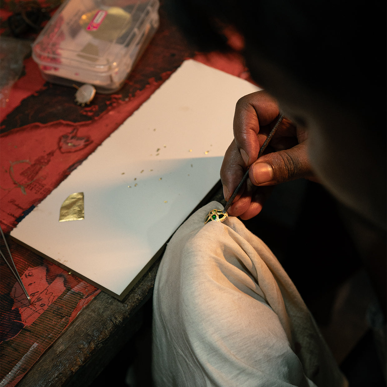 Hands at a bench seen over a dark shoulder: a fine tool against a gold-faced openwork piece with green inserts, a leaf of gold on white board.