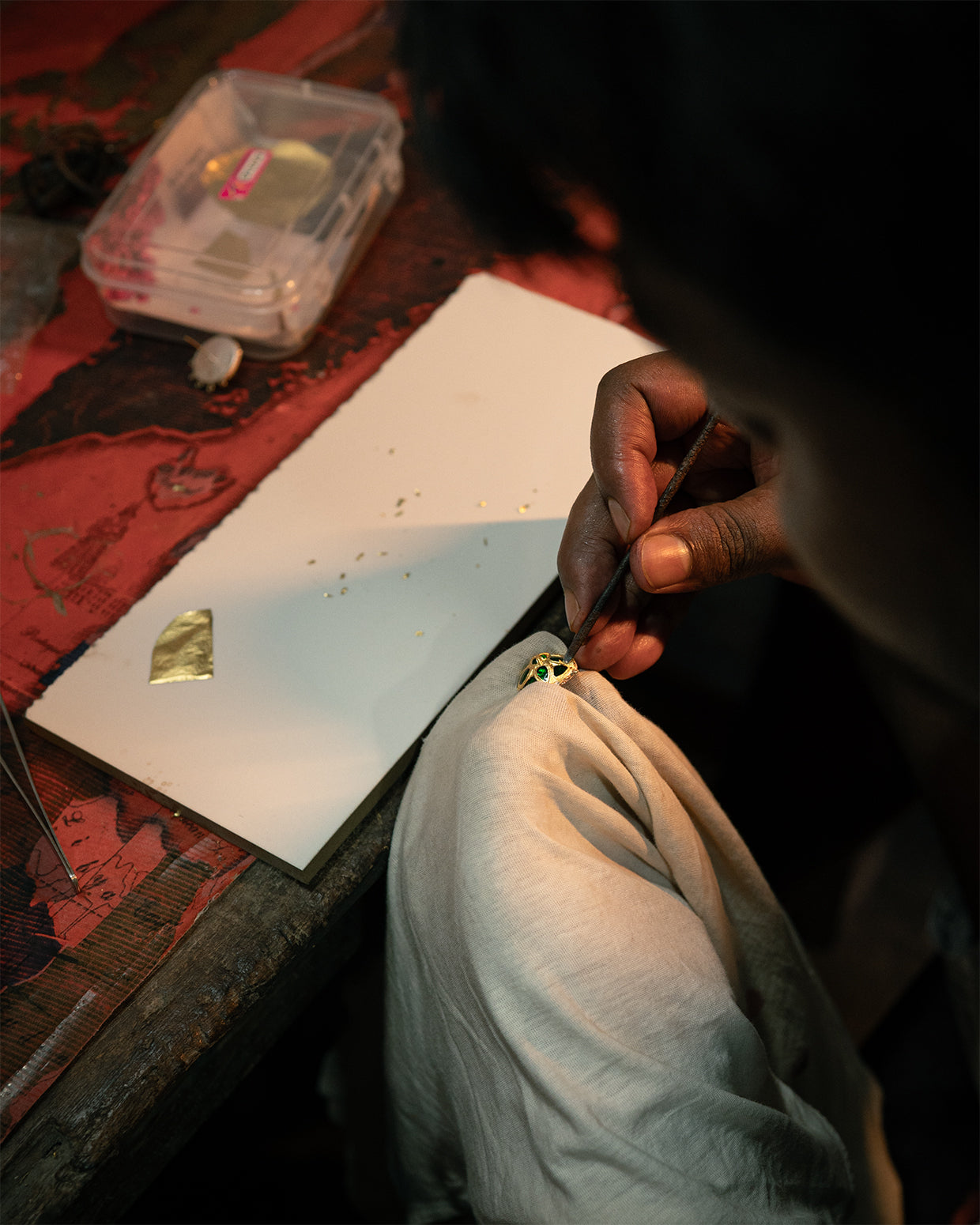 Hands at a bench seen over a dark shoulder: a fine tool against a gold-faced openwork piece with green inserts, a leaf of gold on white board.