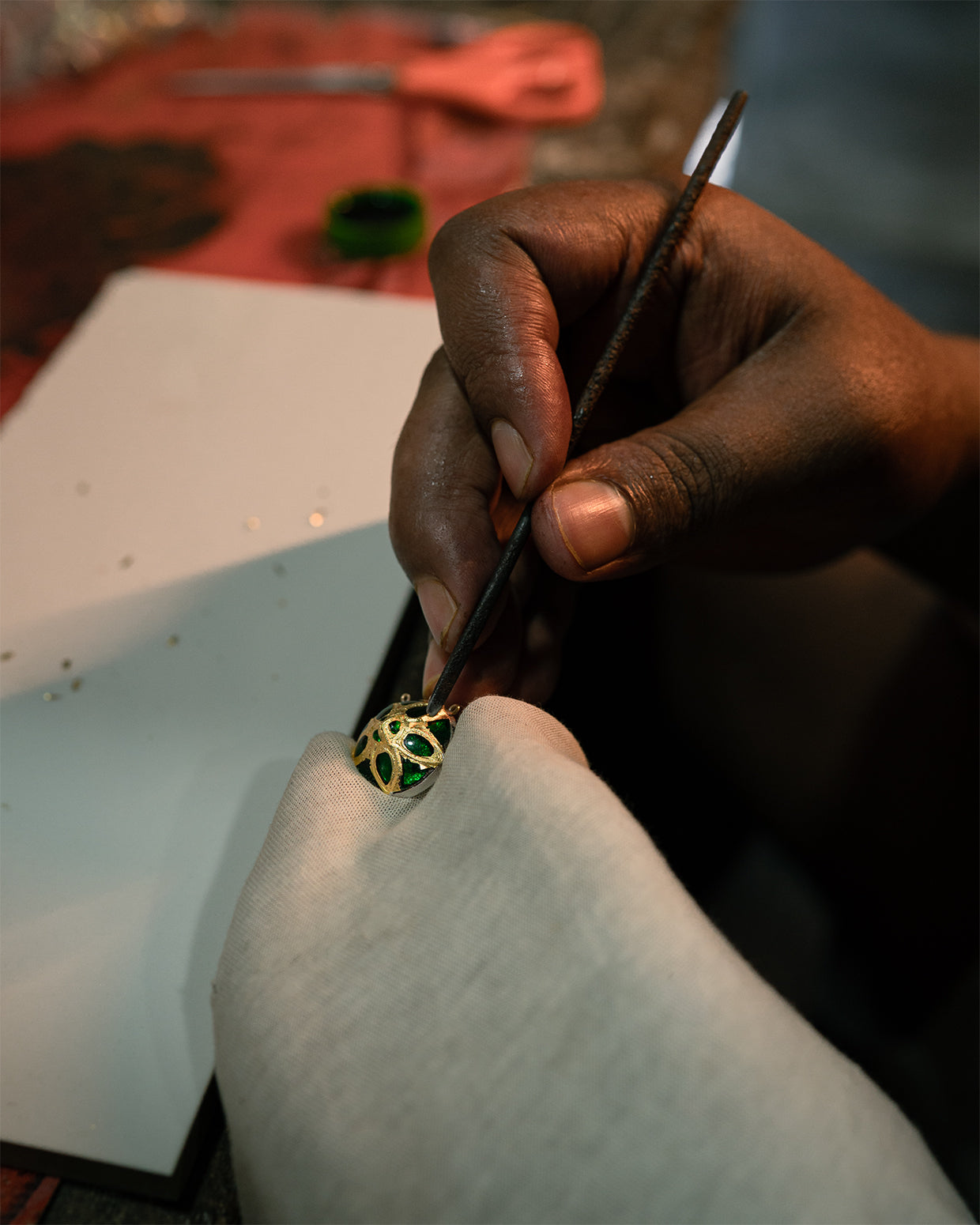 A domed openwork piece close up: gold-faced cells each holding a glossy green teardrop, silver-coloured metal showing below, a tool tip at its edge.