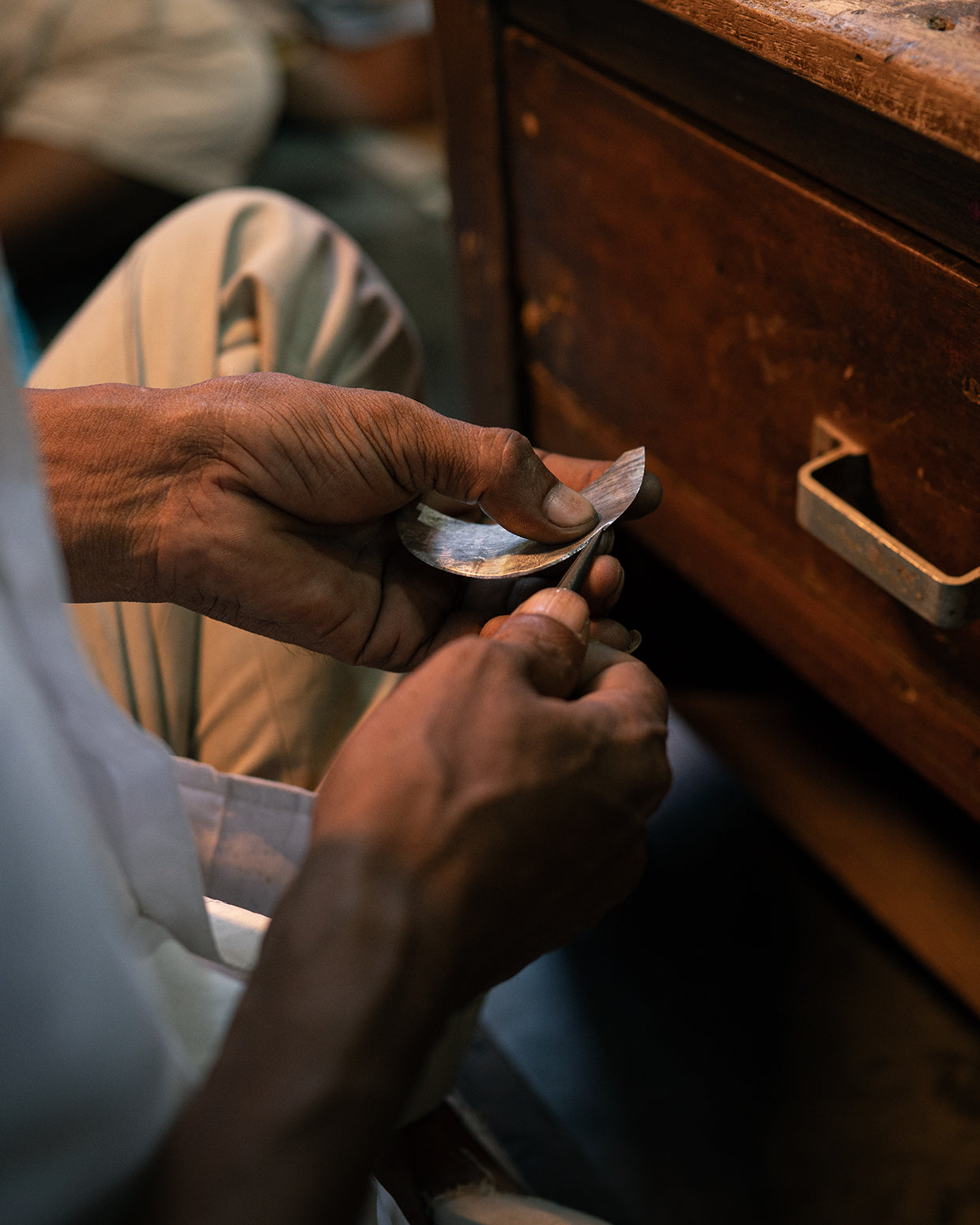 Two weathered hands hold a thin crescent of grey metal and run a narrow wooden-handled tool along its inside curve, beside an old wooden drawer.