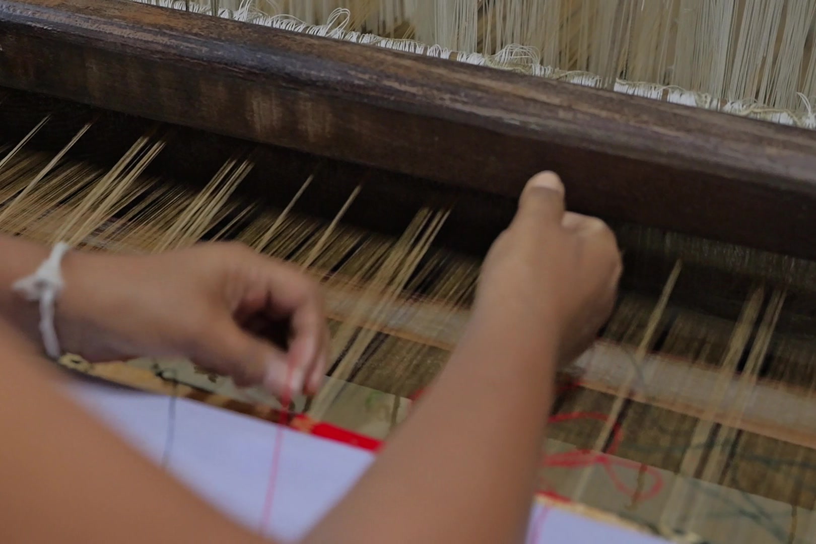 Two hands at the warp of a loom, fingertips parting golden threads above a red motif just woven at the edge of the cloth.