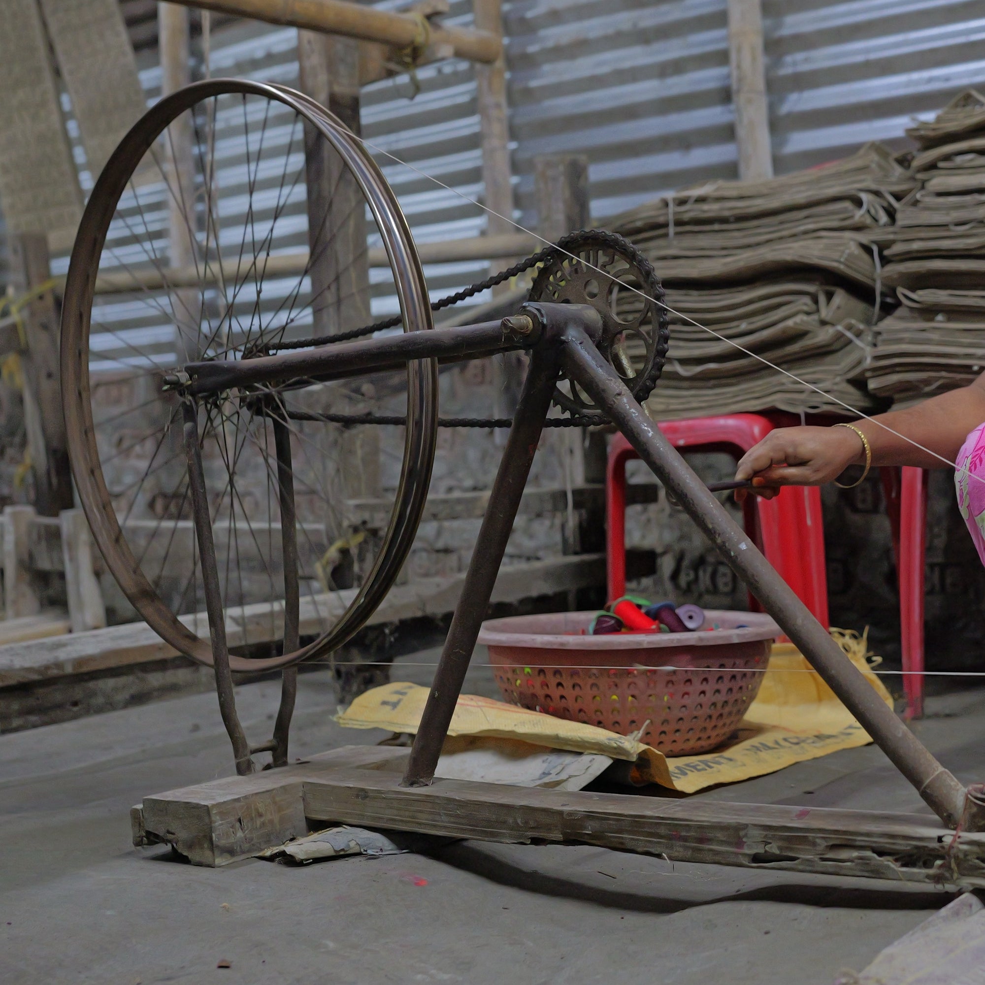 A winding wheel made from a bicycle rim, frame, chainring and chain on a wooden base, a hand on its crank, and a basket of wound pirns in red, green, maroon and blue on a sack beside it.