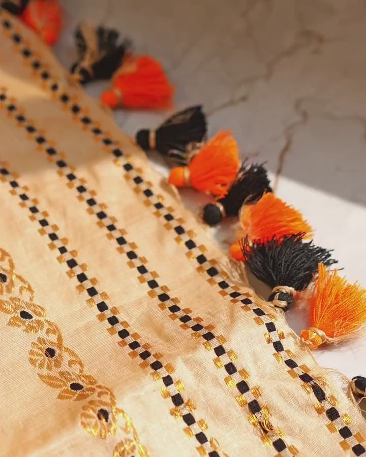 The sador end on a sunlit floor, drifting: a vine of gold flowers with black centres, tiny black-and-gold checks, and orange and black tassels.