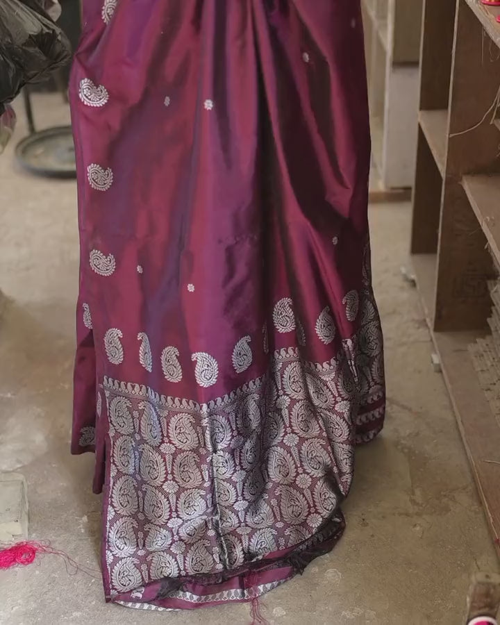 The lower half of the saree close and moving slightly: silver-white paisleys on wine and the dense paisley band at the hem, shelves of reels beside it.