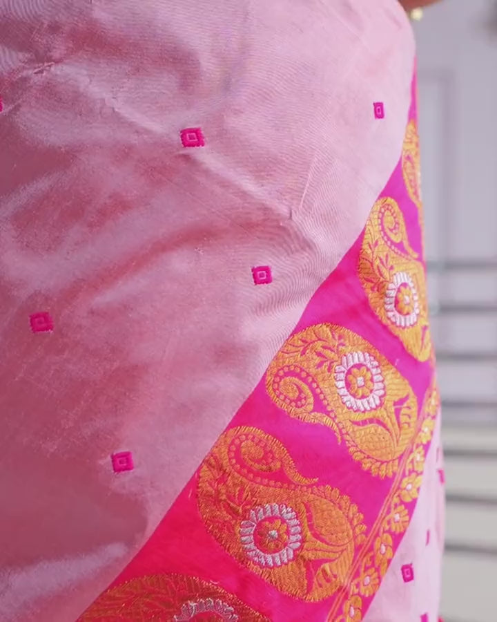 The rose-pink sador shifting over the magenta mekhela's band of golden-orange paisleys; the weave turns between pink and silvery lilac.