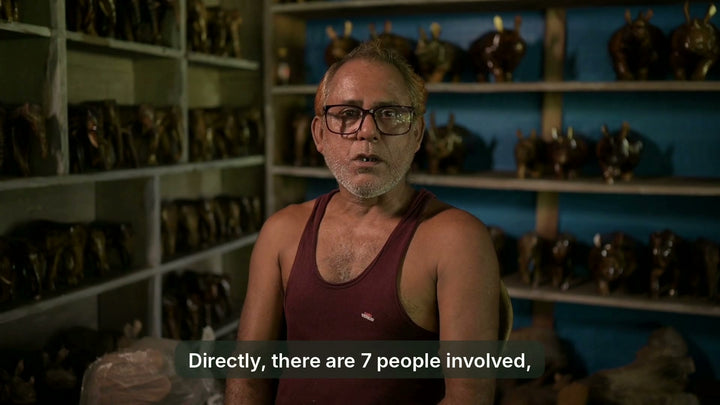 Ghanadaj Chetry in glasses and a maroon vest before shelves of carved rhinos and elephants, speaking about the people who work with him, the Kaziranga rhino and the woods he carves, with English subtitles a phrase at a time. 31 seconds.