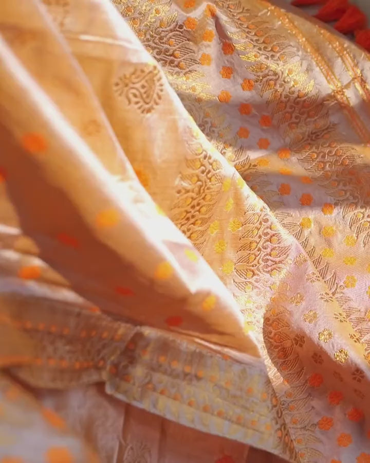 Looking down at the seated lap: sand-beige pleats with saffron and gold florets, the zari border across the top and saffron tassels.