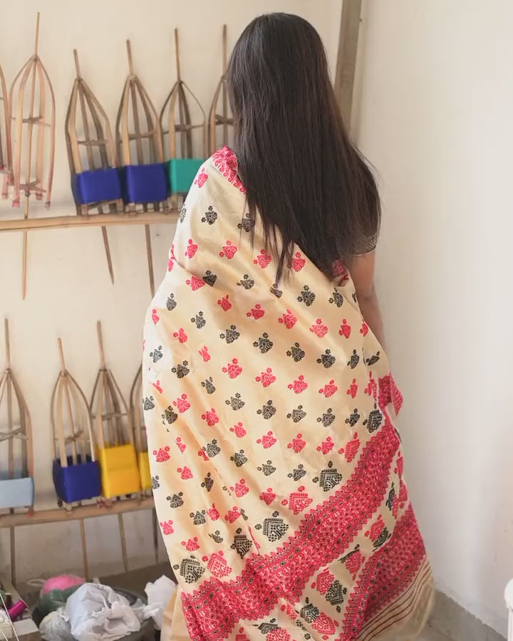 From behind beside the yarn reels, the pallu spread across the shoulders and lifted at its edge: red and black buti on cream, the red scroll border and the pallu’s red bands.