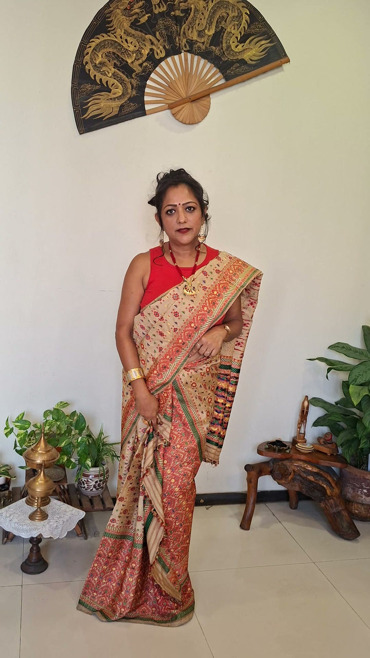 A woman in a red sleeveless blouse puts on a cream and red mekhela sador step by step: the mekhela pleated and tucked in at the waist, then the sador tucked in, taken round and over the left shoulder. She ends by showing the gold jewellery she wears with it.