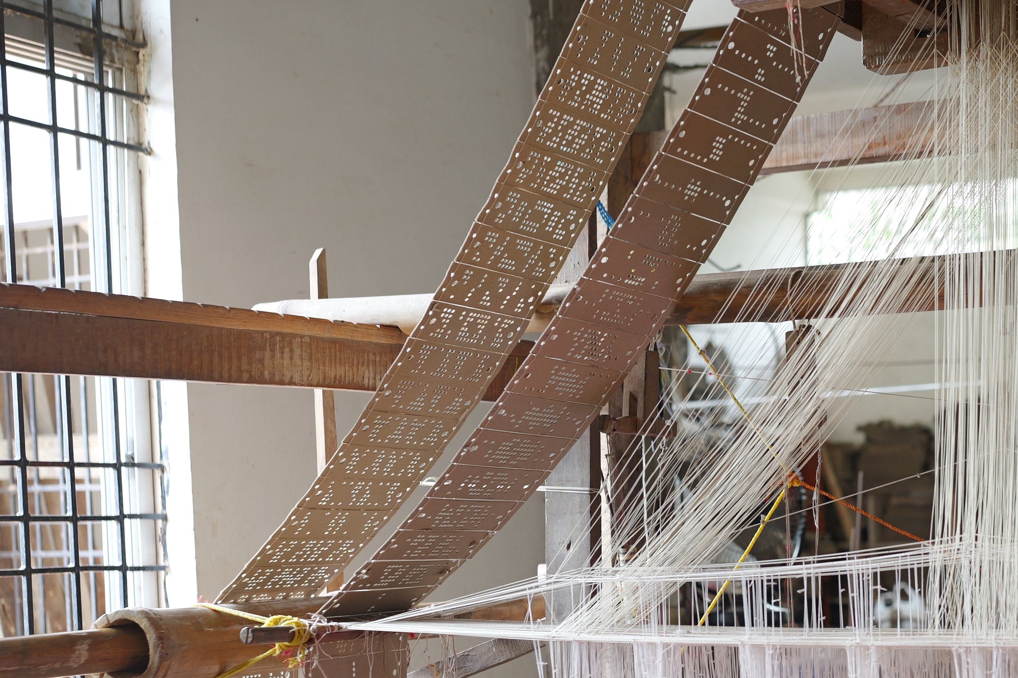 Two long runs of laced punched cards hanging beside a loom, white harness cords converging upward to their right, a barred window behind.