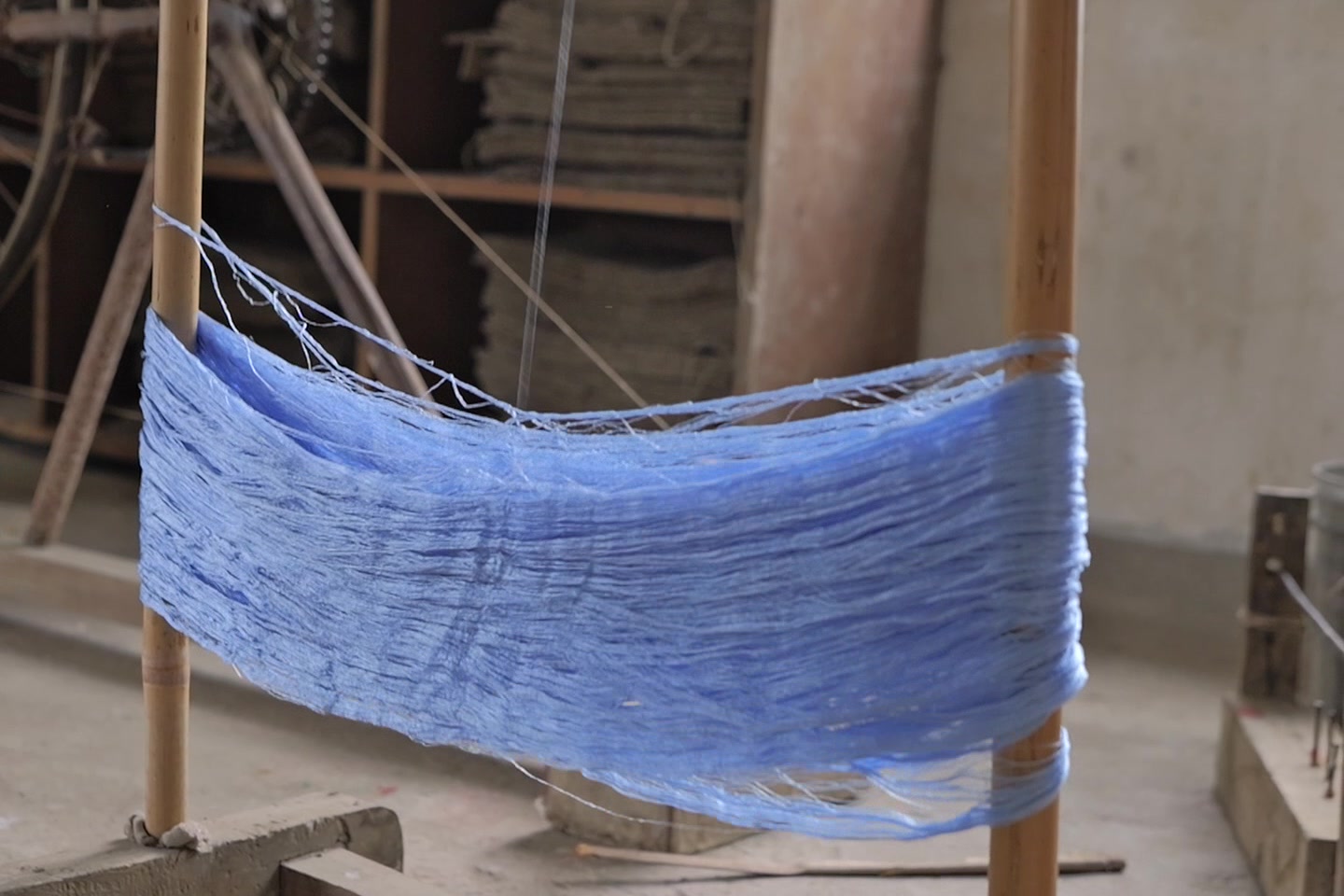 A blue hank stretched between two wooden uprights on a heavy base, a winding wheel and shelves of folded sacking behind it.