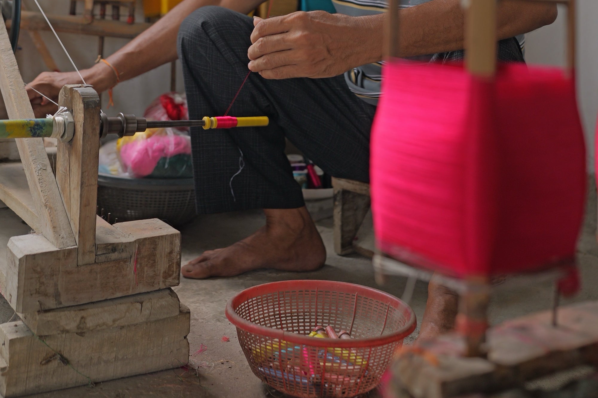 One hand at the wheel of a spindle and the other guiding a magenta thread onto a yellow pirn, magenta yarn on bamboo reels in the foreground and a basket of wound pirns on the floor.