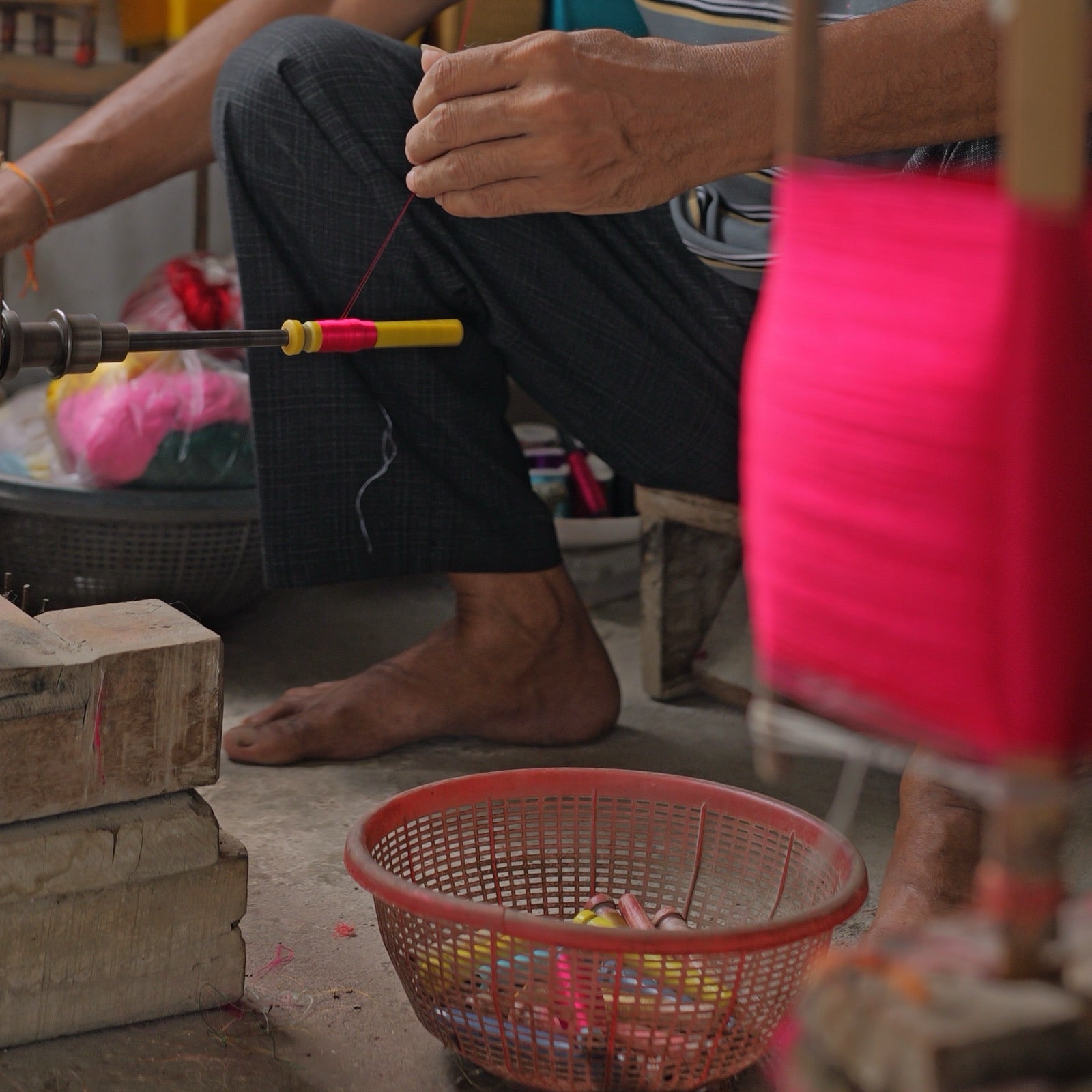 One hand at the wheel of a spindle and the other guiding a magenta thread onto a yellow pirn, magenta yarn on bamboo reels in the foreground and a basket of wound pirns on the floor.
