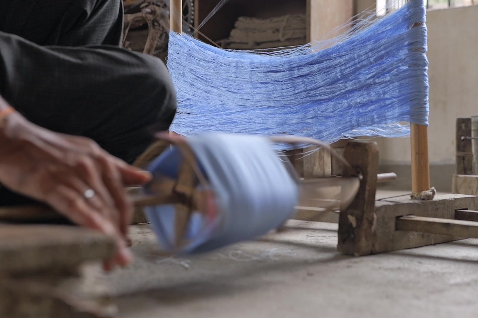 A bamboo cage reel blurred with motion in a wooden floor stand, filling with light-blue yarn from a hank stretched on uprights behind it; a hand with a ring turns it.