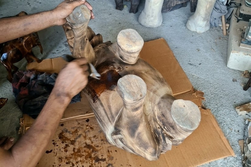 Two forearms over an upturned carved elephant, legs in the air; one hand holds a leg, the other brushes on a dark coat, drips spotting the cardboard.