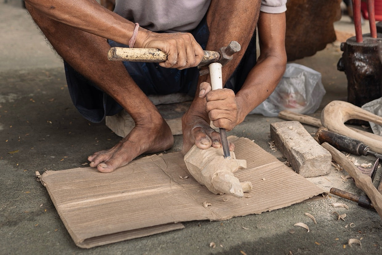A steel hammer strikes a chisel with a white plastic-pipe handle into a pale roughed-out figure held by a bare foot on cardboard, shavings around it.