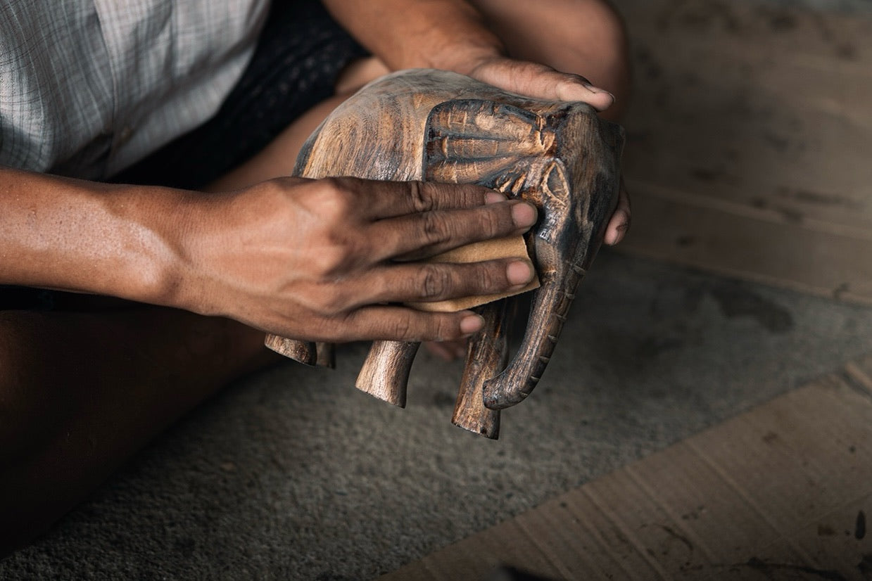 Two hands hold a small carved elephant, one pressing a folded square of abrasive paper against its side; a checked shirt and a cardboard sheet behind.