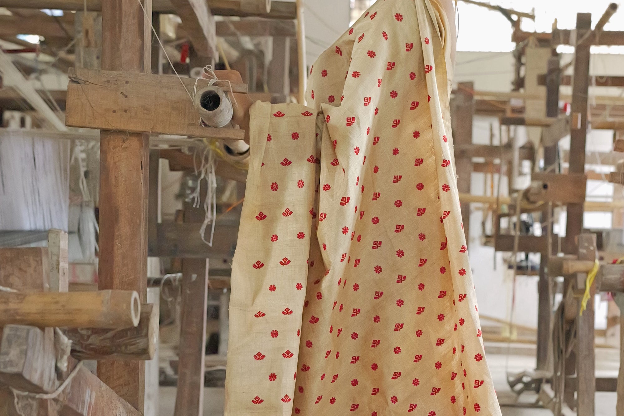 A wooden handloom in a weaving shed, a pirn of thread resting on its frame and a cream Muga cloth scattered with red motifs hanging over it.