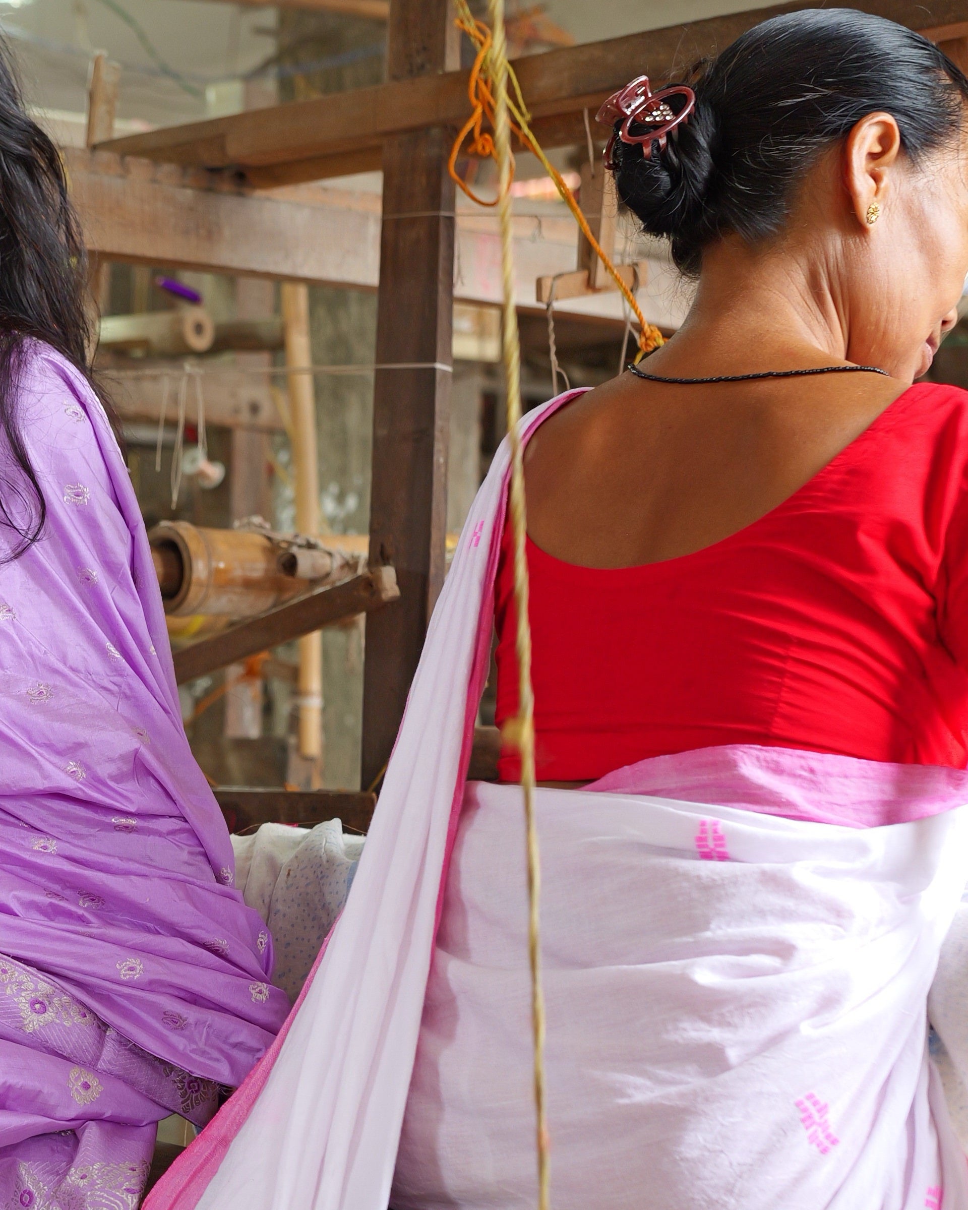 A weaver seated at her loom, seen from behind, a bamboo frame and hanging warp threads in front of her.