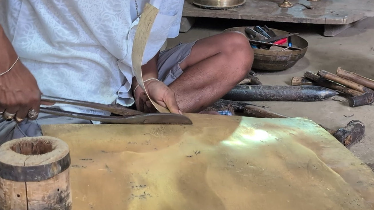 Hands with rings work long iron shears along a yellow metal sheet on the floor, a cut strip curling up; hammers and an iron stake lie behind.