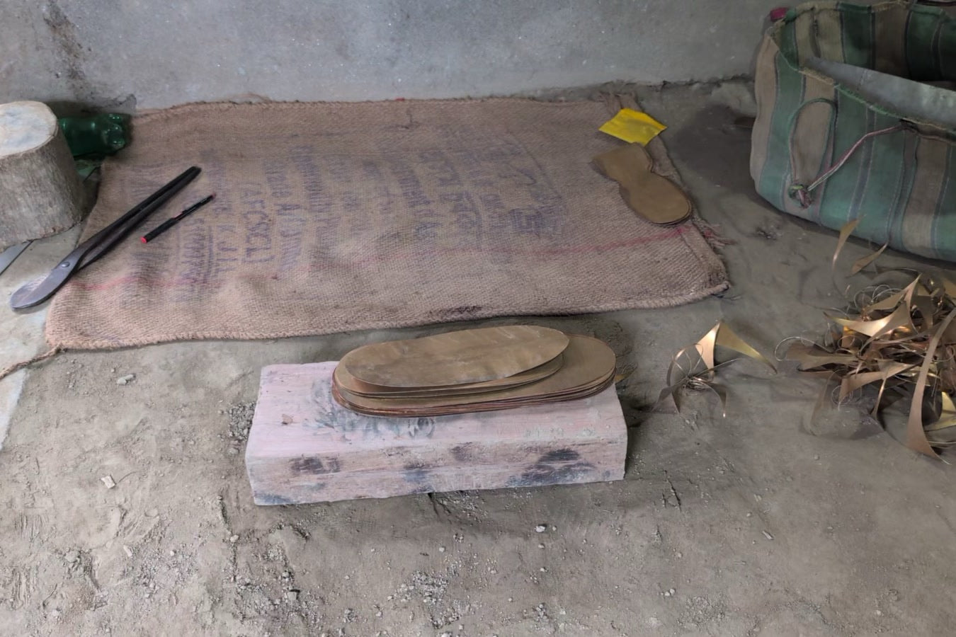 A stack of flat oval metal blanks on a pink-painted block, long hand shears and a marker on a jute sack behind, curled metal offcuts at the right.