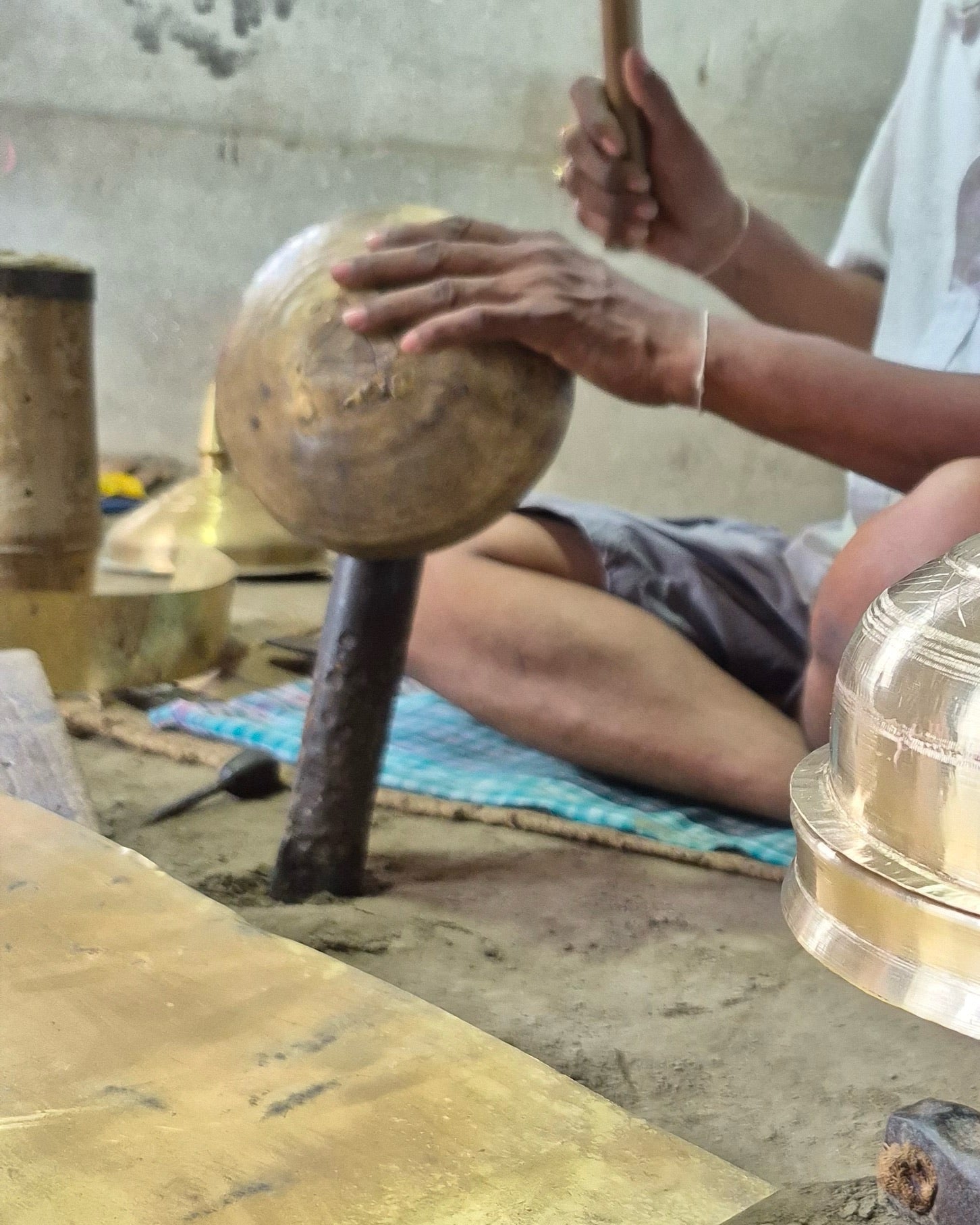 A hand holding a rounded, dimpled metal dome over an iron stake set in an earth floor, the handle of a hammer in the other hand.