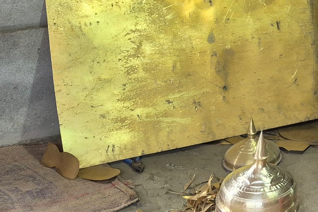 A large sheet of yellow metal leaning on a concrete wall, flat offcuts on the floor at its foot and two polished domes with pointed tips in front.