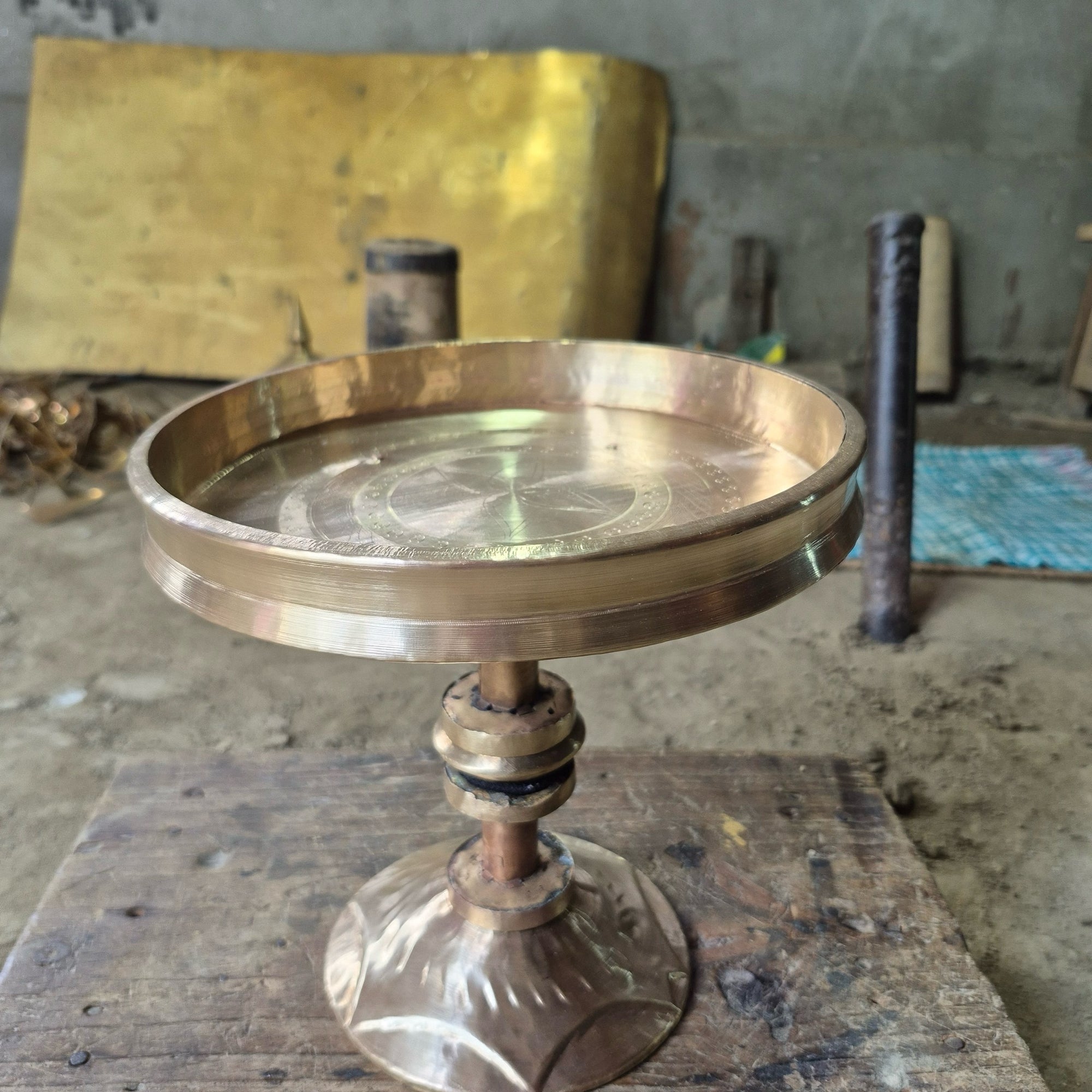 A lidless metal tray on a copper-coloured tube stem with collars and a lobed, flared foot, standing on a scarred wooden bench.